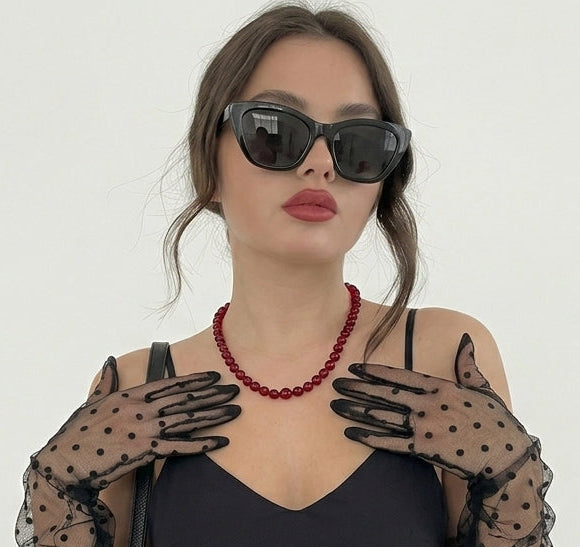 Woman wearing black sunglasses, a black dress, and black polka dot gloves on a white background carries a red amber necklace for women